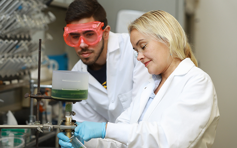 professor and student at a lab station wearing lab coats and goggles observe a beaker that is connected to a burner