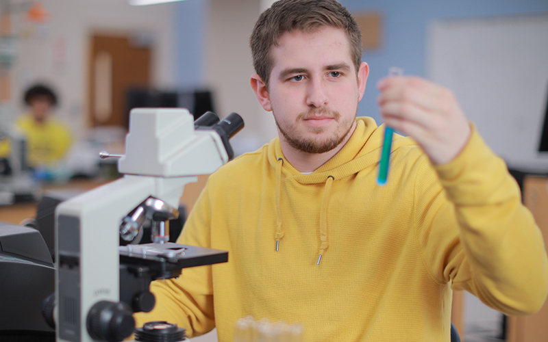 student in a classroom sitting infront of a microscope holding up a test tube full of liquid