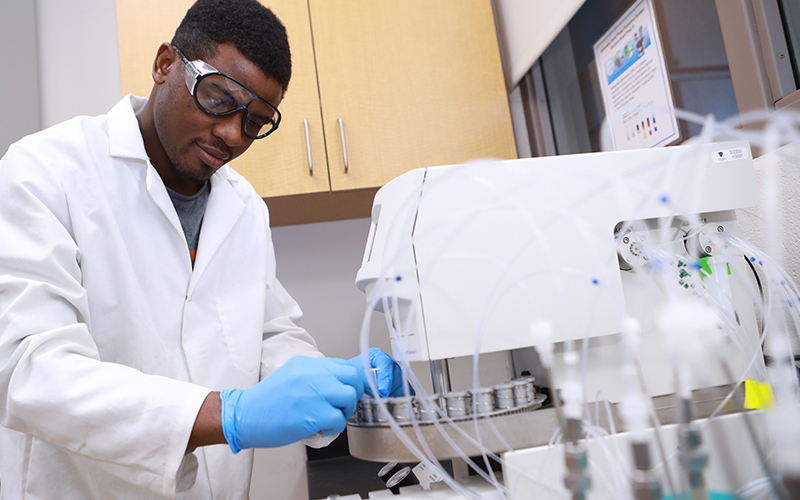 man wearing a white lab coat and goggles assembles and adjust small tests tubes inserted in a machine