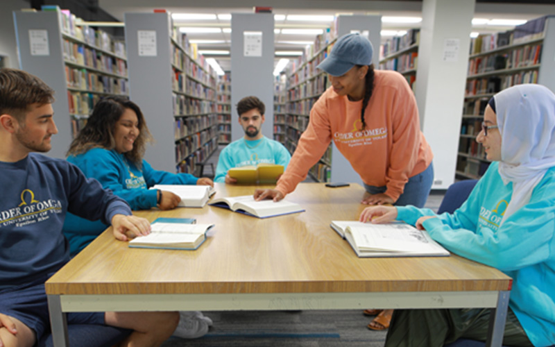 group of students sit at table together in the library looking at their textbook