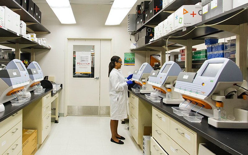 woman in a white lab coat and goggles stands in front of a row of lab machinery gathering measurements 