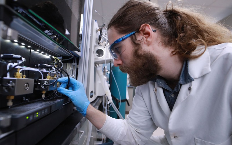 man in white lab coat and goggles adjusts lab machinery 