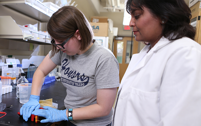 professor in a white lab coat observes as student wearing gloves works with material at a lab station