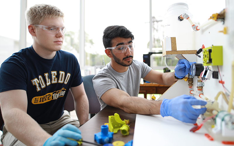 two students working at a lab station wearing goggles and gloves 