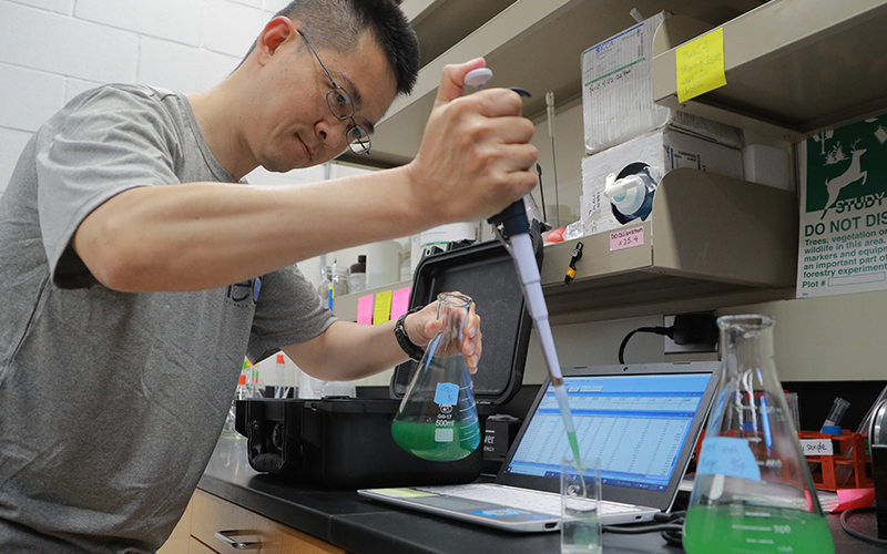 Man in a lab using a pipette to transfer liquid from a large flask to a small glass beaker