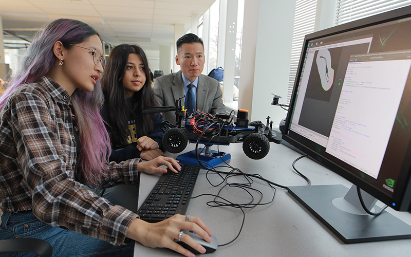 two students program a miniture electric car at a computer station with the assistance of their professor