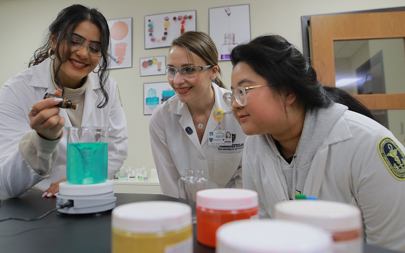 three students in lab coats and goggles watch as one student adds drops into a beaker