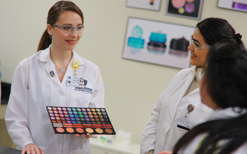 student in lab coat and goggles presents makeup palette to two other classmates