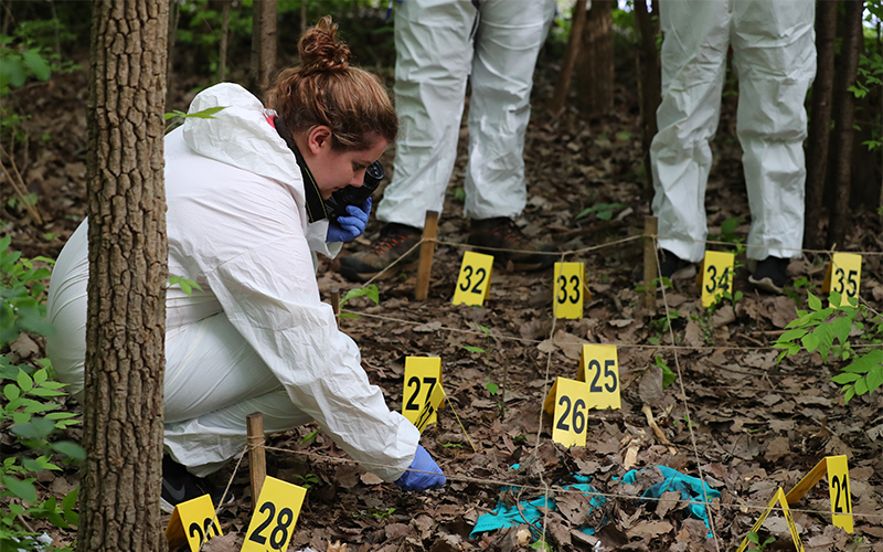 Girl in a white hazard uniform in the woods with a camera inspecting the materials in front of her