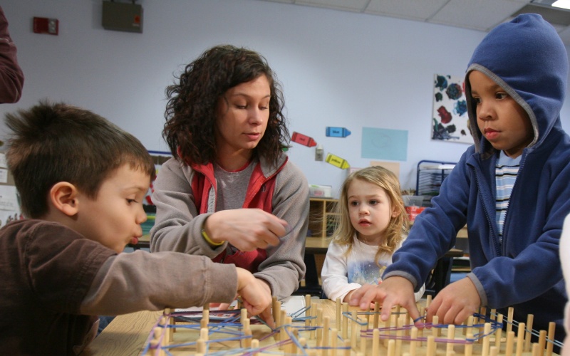 a teacher sits at a table with three young students during an activity of looping rubber bands onto a board with small wooden poles