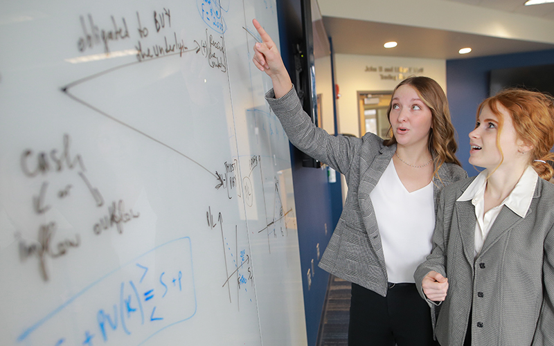 two students point to and observe data written on a white board