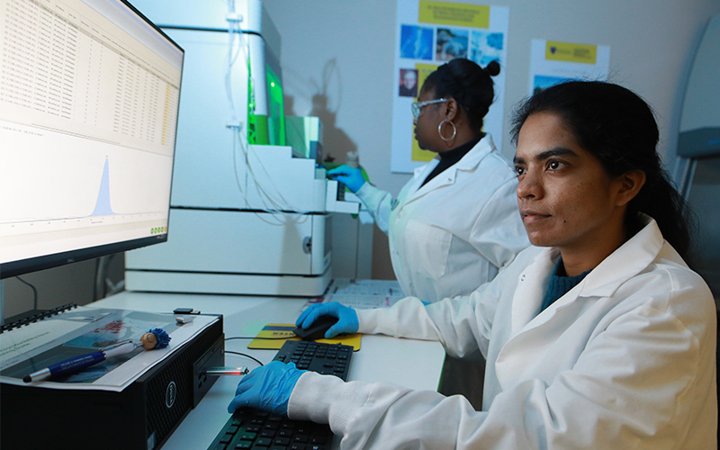 Woman in a labcoat on a computer looking at data tables another woman in the background in a labcoat looking through cabnets 