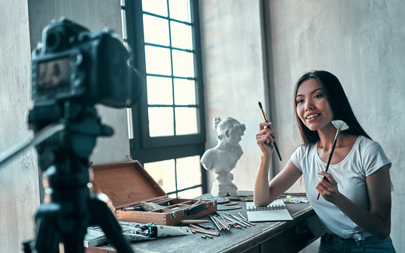 woman sits in front of camera and films video talking while holding two different paint brushes