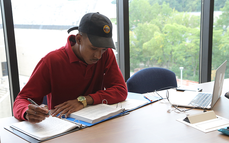 student sitting at a table in the library with laptop and textbook open takes notes