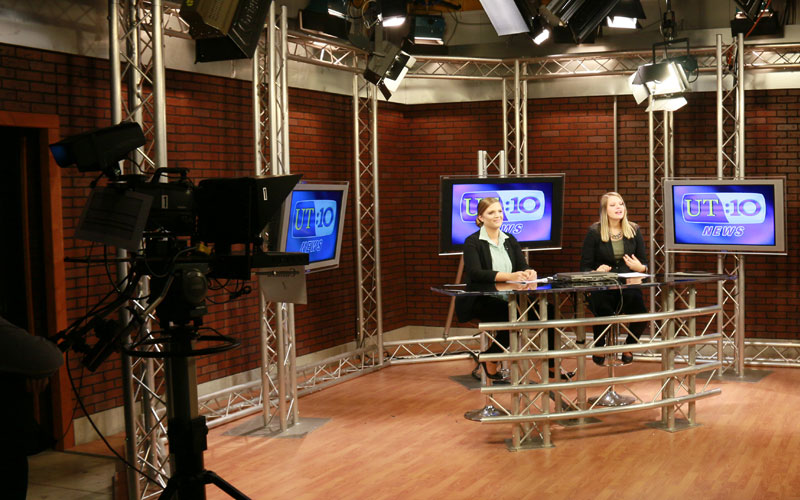 behind the cameras perspective of a broadcast featuring two hosts sitting at a table on stage in front of the film cameras
