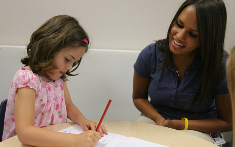 teacher sits next to young student writing at a table 