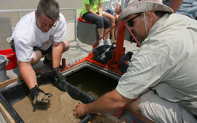 two scienists on a boat crouch down over a tub of muddy water and sift for samples