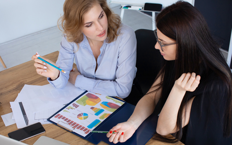 two women sit at a table observing a portfolio containing pie charts and graphs