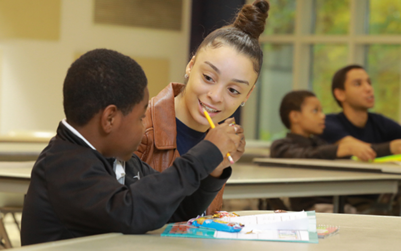 a woman sits besides and talks to a young child at a table who's holding a pencil 