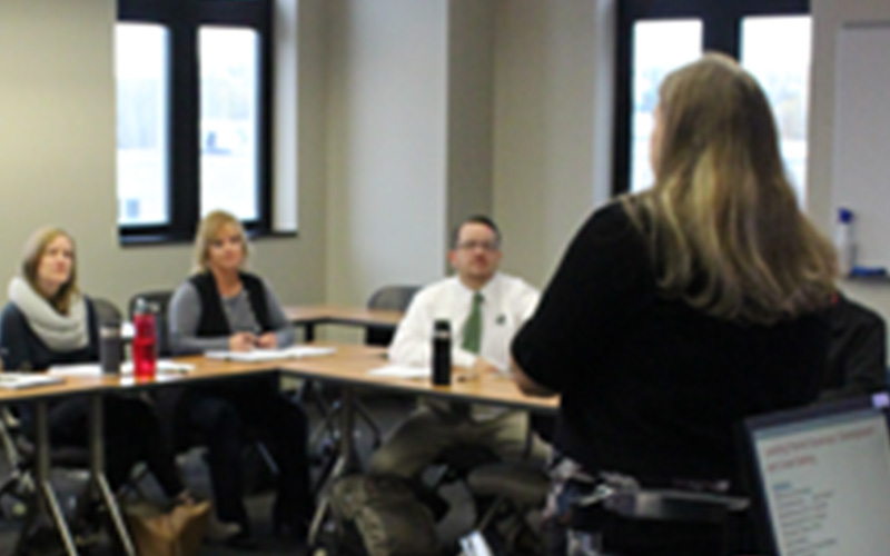 woman presents in front of three students that are sitting at tables in the classroom