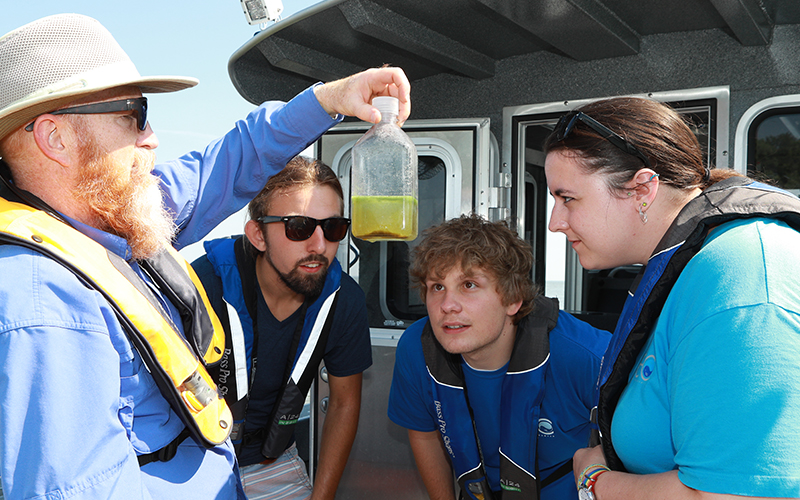 4 people on a boat all wearing life jackets the professor is holding up a bottle of liquid while the three students closly observe the bottle the professor is holding