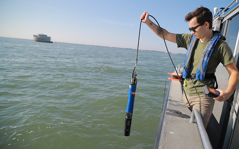 Man standing on a boat wearing a life jacket holding an electronic device over the side of the boat above the water