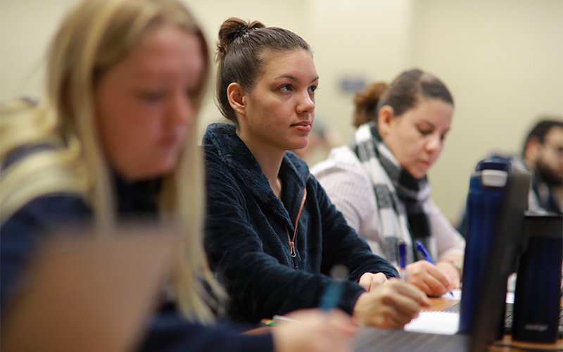 a row of students seated at a lecture hall table take notes in their notebooks