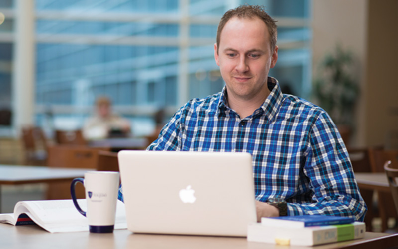 man sitting at table with open laptop and textbook