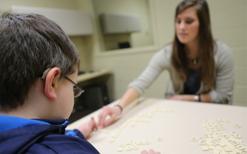a woman sitting across a young child at a table helps piece together words using small tiles with letters 