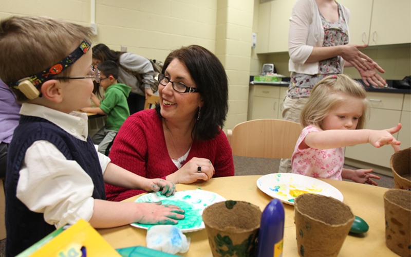 a woman crouches down at a table with two young children using paint for a craft