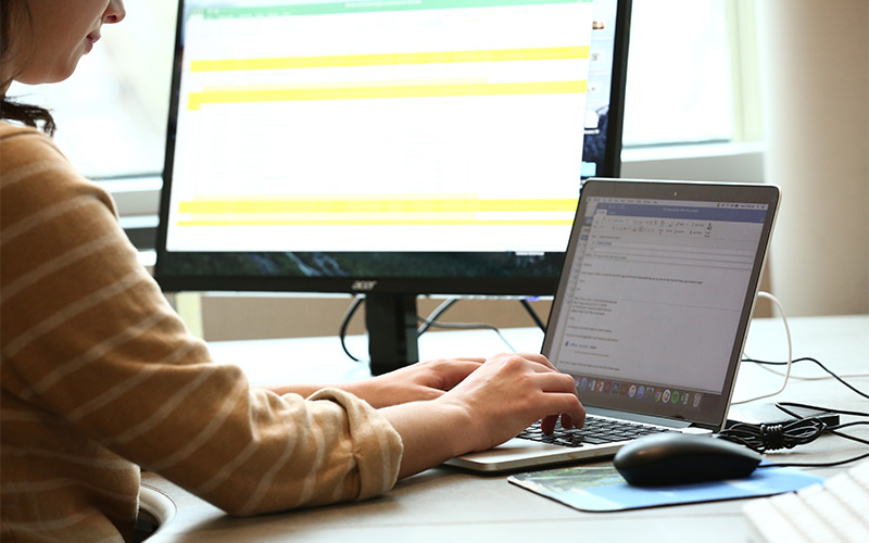 woman sits in front of two computers and types message on laptop