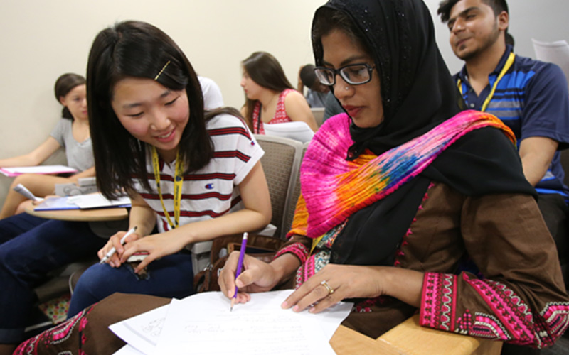 two students sit next to each other in desks and discuss their assignment