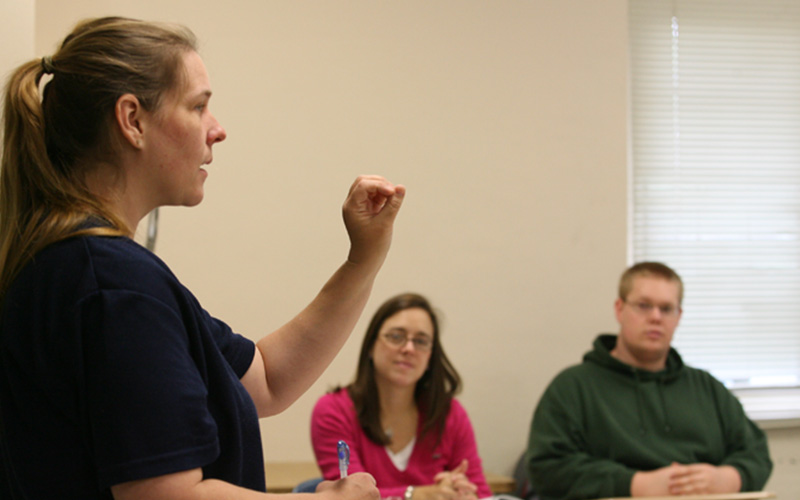 professor stands and teaches in front of class, two students sit next to each other at a table listening to the lecture