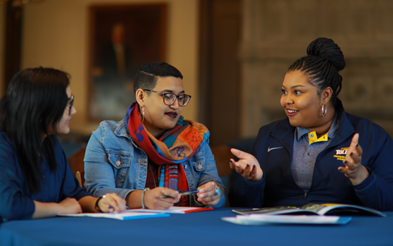 Three students sit at a table with open notes and textbooks discussing the material 