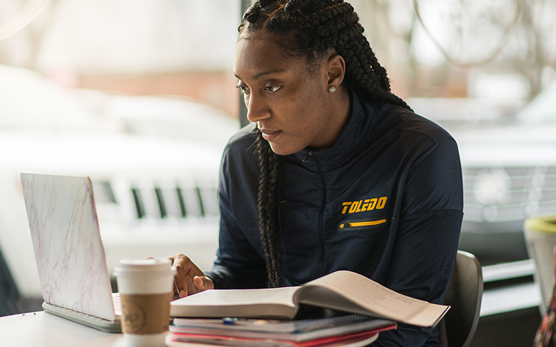 woman observes laptop screen at a table with open notes and textbook