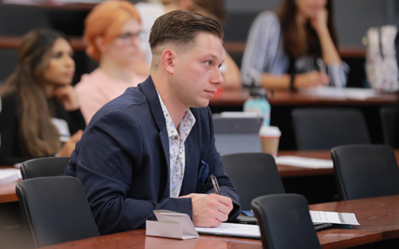 student sits at table and intently listens to lecture while taking notes 