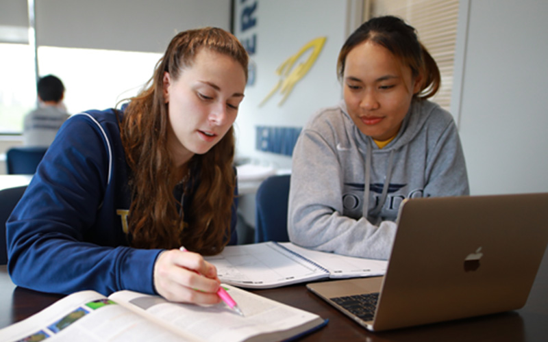 two students sit together looking at an open textbook 