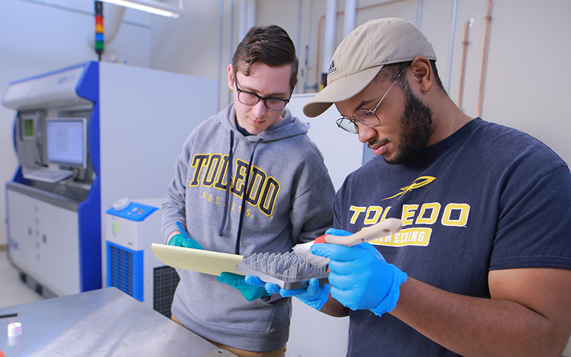 one student takes notes on a notepad while the other students holds a brush to a piece of metal with spikes
