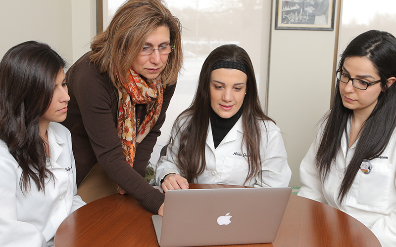 three women in labcoats sitting at a table gatherd around a 4th women who is standing up controlling the laptop while the other 3 women observe