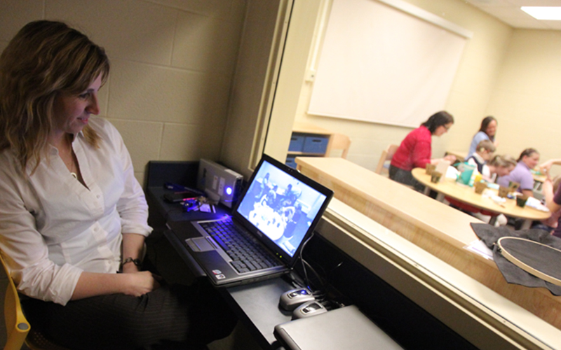 a woman sitting behind a window in an observation room watches the other adult attendants interact with the young students in the room on the other side
