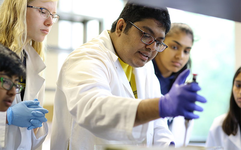 a group of students wearing white lab coats and gloves observe as one student dispenses a liquid from a dropper
