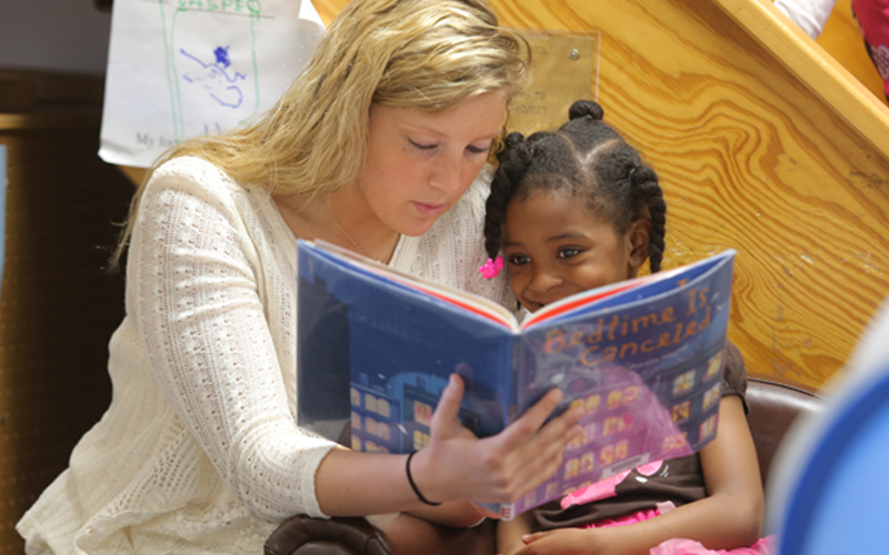 teacher sits next to a young student and reads from a storybook 