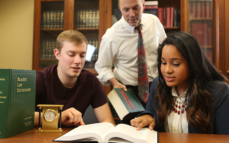 two students sit at a table with an open textbook with one student pointing out to a section of text