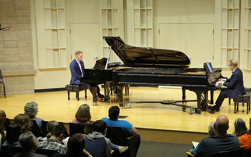 two men on piano perform on stage in a concert hall in front of an audience