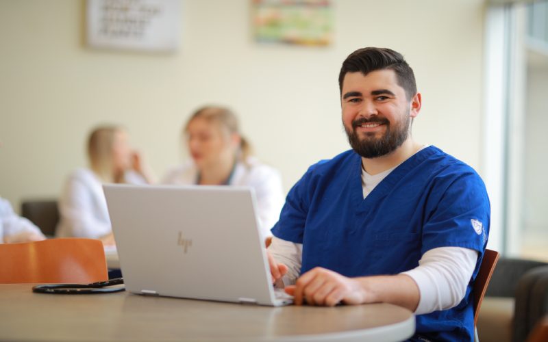 a man wering blue nursing scrubs sits at a table with an open laptop