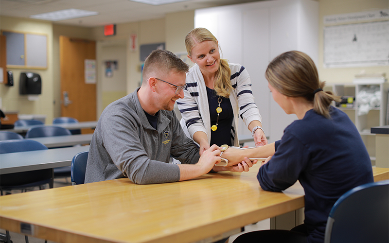 a man sits across from a woman wearing an arm device gathering medical measurements in a classroom lab setting 