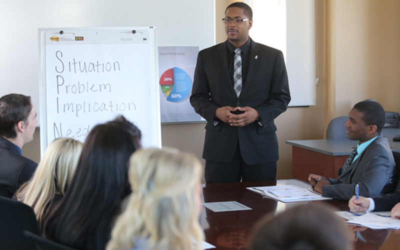 man stands in front of group at a conference table and presents information on white board