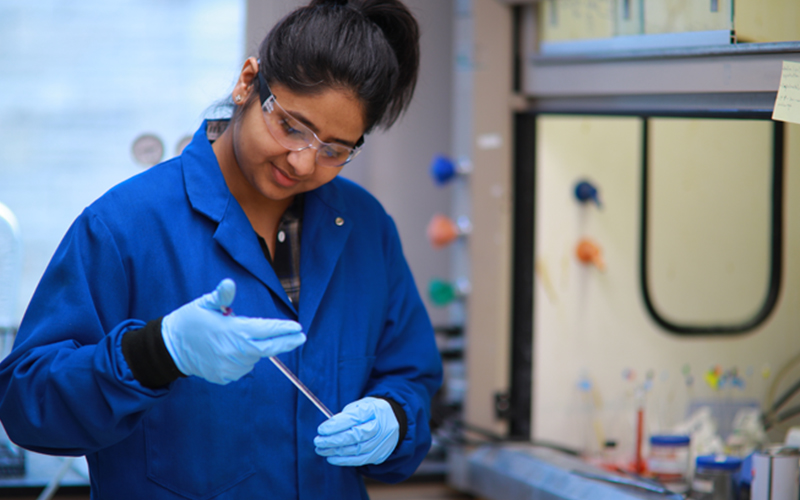 woman wearing a blue coat and goggles holds and observes a test tube