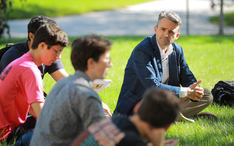 a professor listens in on group discussion during a lecture taking place outside in a grassy patch