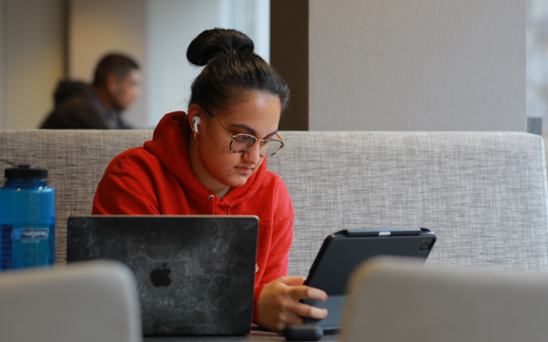 student studies in the library at a table, sitting in front of an open laptop and tablet